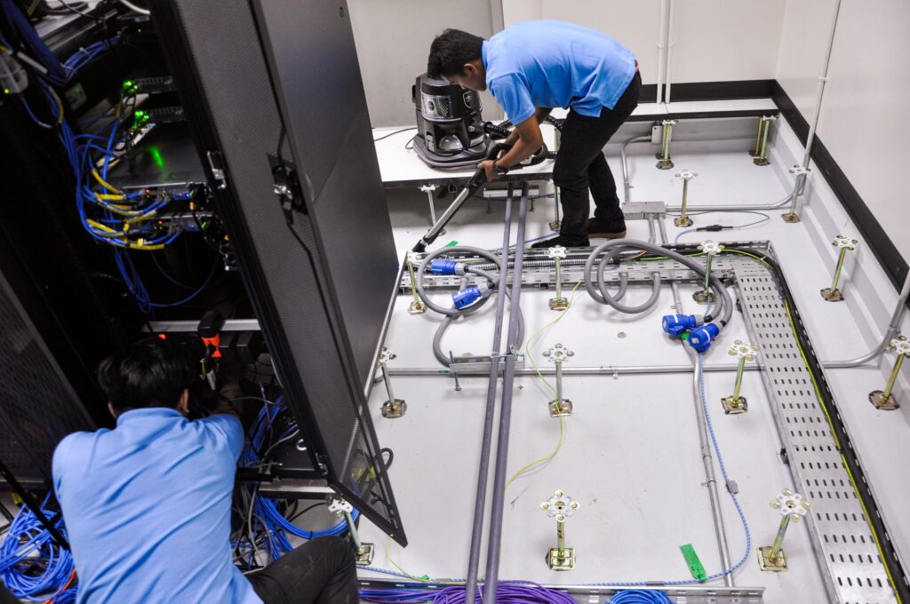 This photo shows specialized cleaning professionals using HEPA-filtered vacuums to safely remove sub-floor contaminants without disrupting active server equipment.