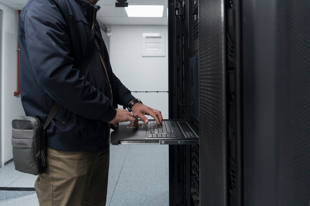 Data center technician performing maintenance inside a server rack aisle with raised floor access and structured cabling in a high-density facility.